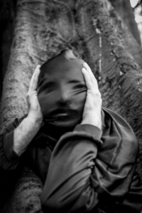 Black and white photo of a hooded figure with blurred face leaning against a tree