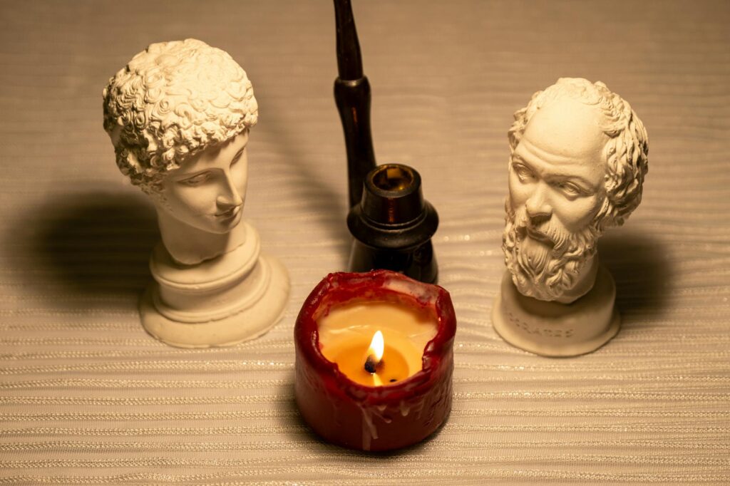 Two classical busts beside a lit candle and antique pipe on a textured surface